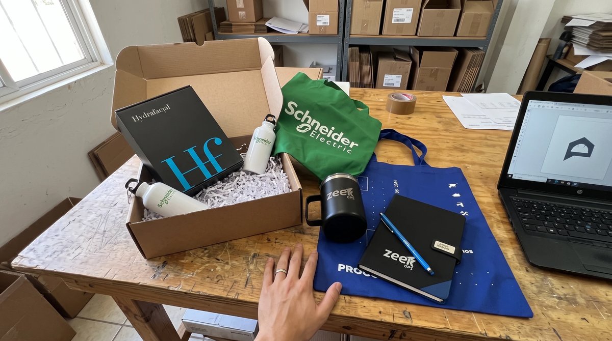 Caja abierta con distintos artículos promocionales personalizados de varias marcas: botellas, tote bag, taza y libreta con etiqueta de nombre